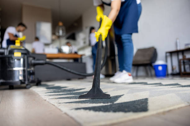 Person vacuuming a geometric-patterned rug in a modern living room, wearing yellow gloves. Background shows blurred figures and furniture.