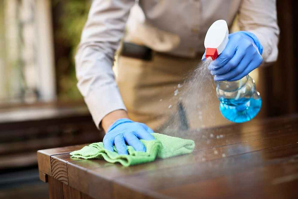 Person cleaning a wooden table with blue spray and green cloth. Wears blue gloves. Indoor setting, focused, tidy atmosphere.