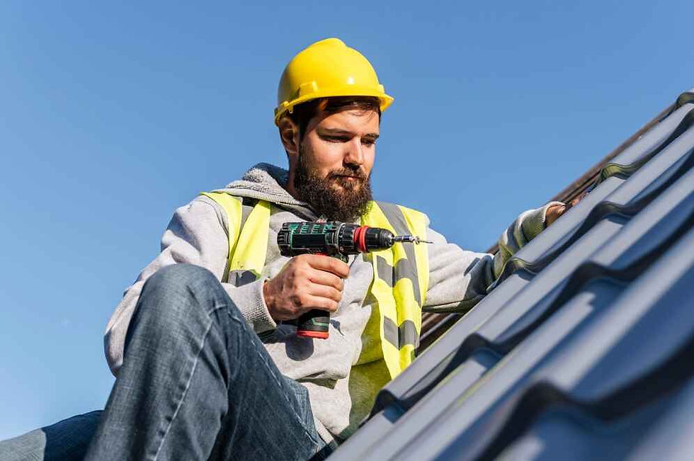 Construction worker in a yellow hard hat and vest uses a power drill on a metal roof under clear blue sky, focused expression.