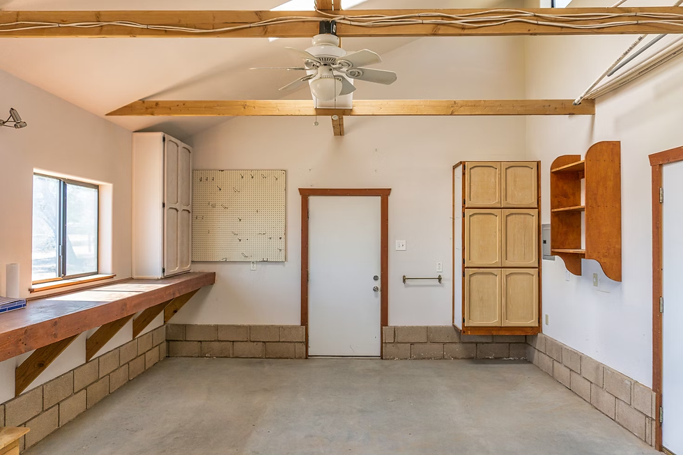 Empty garage interior with beige walls, wooden beams, ceiling fan, pegboard, and cabinets. Sunlight streams through the window.