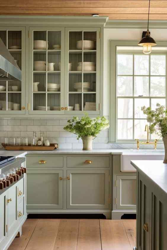 Light green kitchen with glass cabinets, white dishes, and a farmhouse sink. Wood floors, subway tiles, and potted plants add a cozy feel.