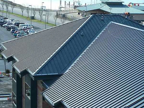 Metal roofs of adjacent buildings with a parking lot and road in the background. Overcast sky; rooftops create geometric patterns.