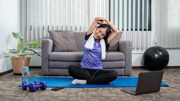 Woman stretching on a blue mat in living room, smiling. Laptop, dumbbells, and water bottle nearby. Gray couch and plants in background.