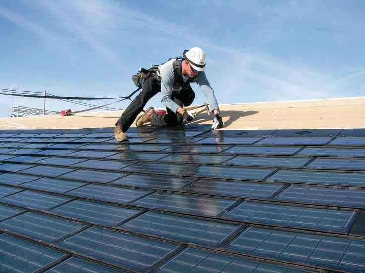 Worker installing solar panels on a roof under clear blue sky, wearing safety gear and helmet, with solar tiles reflecting sunlight.