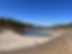 Dry lakebed with scarce water, surrounded by forested hills under clear blue sky. Sandy shoreline curves around the remaining pool.