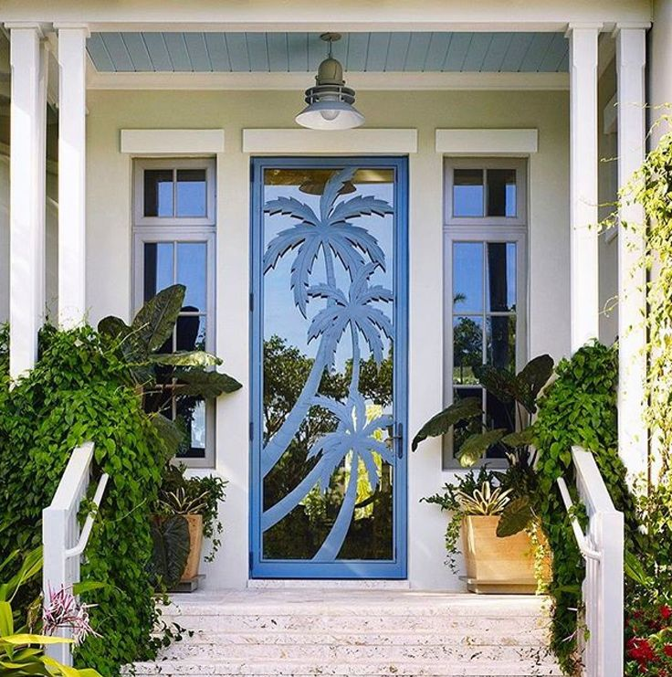 Blue door with palm tree design flanked by potted plants on a porch. White columns and soft lighting create a tropical ambiance.