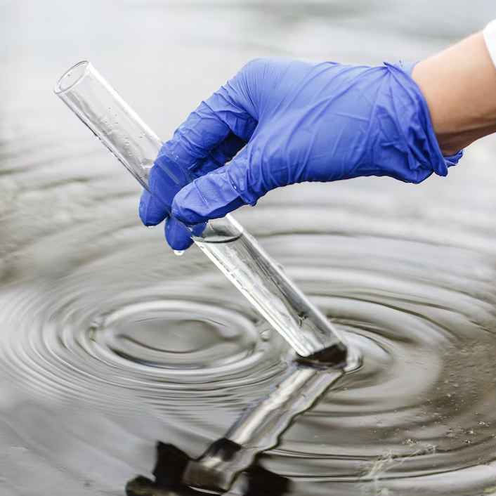 A gloved hand dips a test tube into rippling water, suggesting water sampling. The blue glove contrasts with the water's reflective surface.