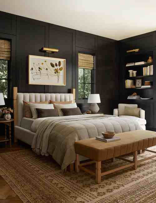 Cozy bedroom with dark walls, large bed with soft gray bedding, framed art above, armchair, shelves with books, and a patterned rug. Warm ambiance.