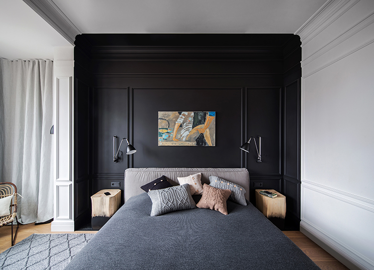 Modern bedroom with a dark accent wall, gray bedding, and multiple pillows. Abstract painting and dual wall lamps above the bed. Cozy mood.