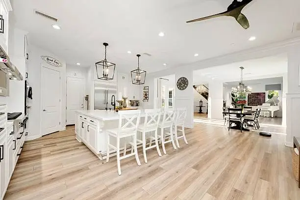 Bright kitchen with white cabinets, island, and chairs. Modern lighting, wood flooring, and wall clock. Dining area visible in the background.