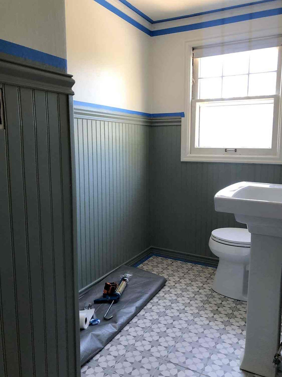 Bathroom with gray wainscoting, geometric tile floor, and blue painter's tape. Tools and paper towel on the floor. Bright window light.