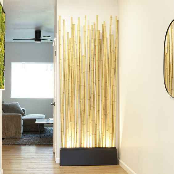 Tall bamboo stalks in a sleek planter, softly lit, decorating a modern hallway with white walls; living room visible in the background.