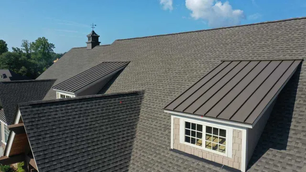 Steep gray shingle roof with dormer windows, metal accents, and a cupola under a blue sky. Green trees are visible in the background.