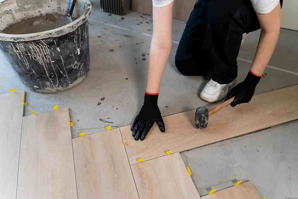 Person installing wood flooring, kneeling with a mallet. Gray bucket with tools nearby. Yellow spacers visible. Casual, focused mood.