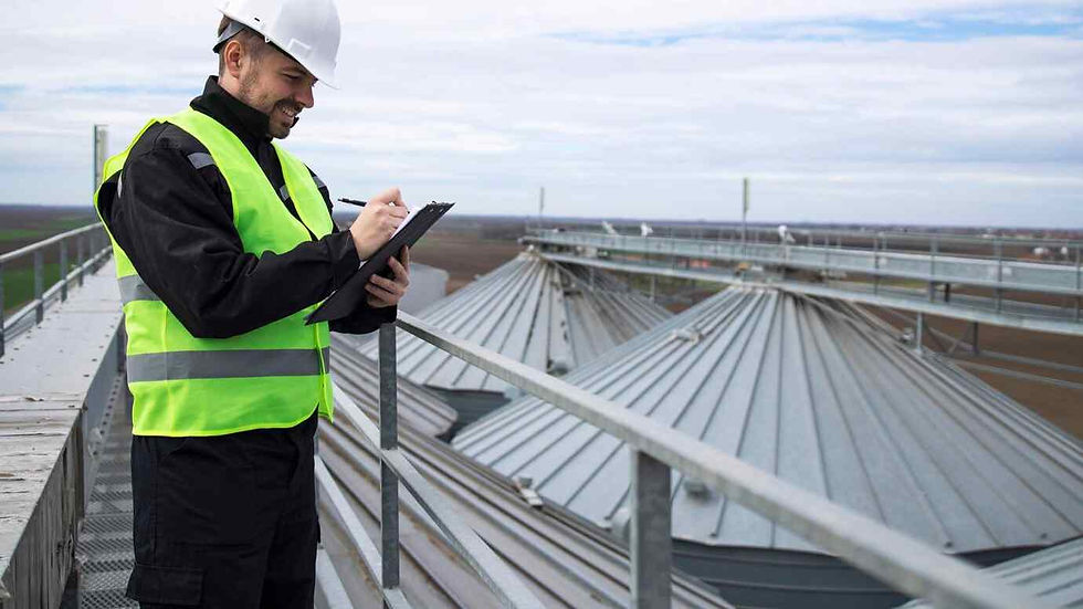 Worker in a yellow vest and white hard hat writes on a clipboard atop a metal walkway, smiling. Silo rooftops and cloudy sky in background.