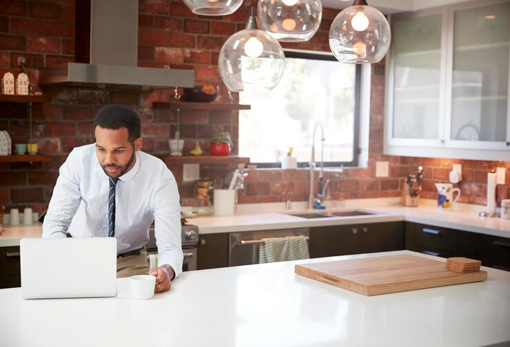 Man in white shirt and tie drinks coffee, works on laptop in modern kitchen with brick walls, pendant lights, and sunlight through window.