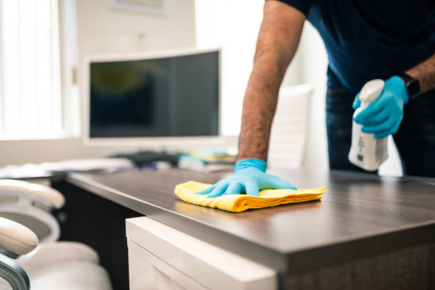 Person cleaning a desk with yellow cloth and spray in an office. Wearing blue gloves. Background shows a computer monitor and chairs.