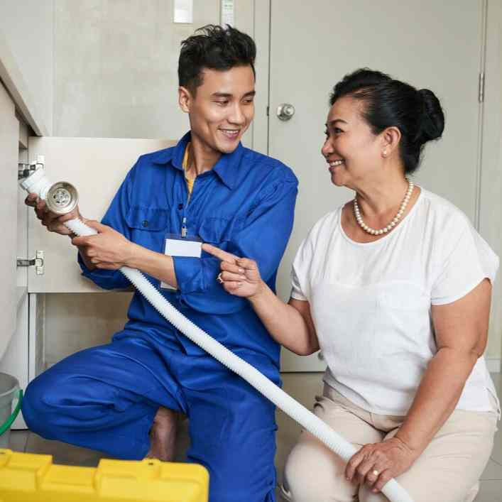 A plumber in blue overalls shows a hose to a smiling woman in a white shirt in a kitchen. They're kneeling by a cabinet, yellow toolbox nearby.