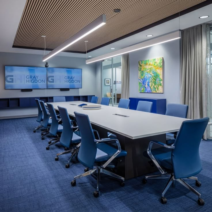 Modern conference room with blue chairs around a long white table. Two screens show "Gray Higdon." Bright artwork and soft lighting create a calm mood.
