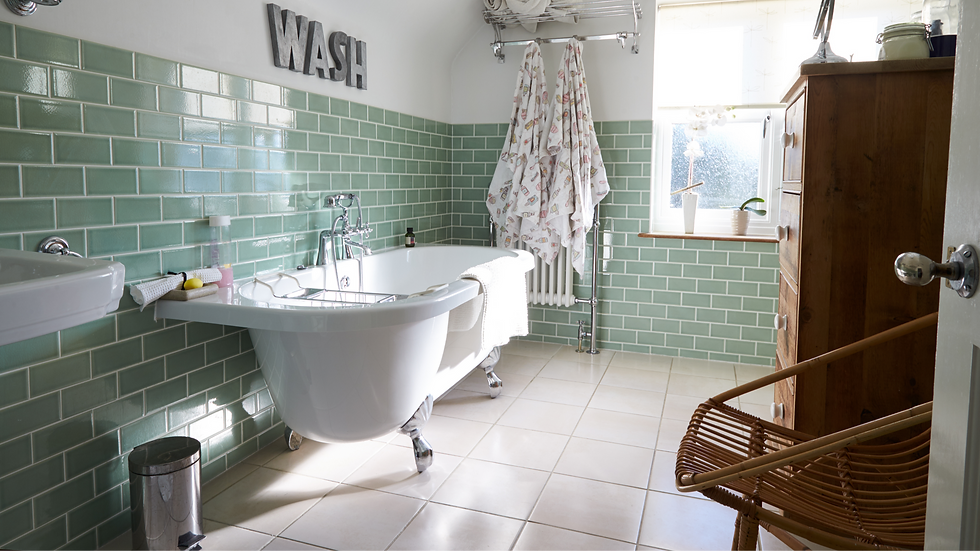 Bright bathroom with a clawfoot tub, green tiles, white sink, and wooden cabinet. "WASH" sign on wall. Sunlit window, cozy ambiance.