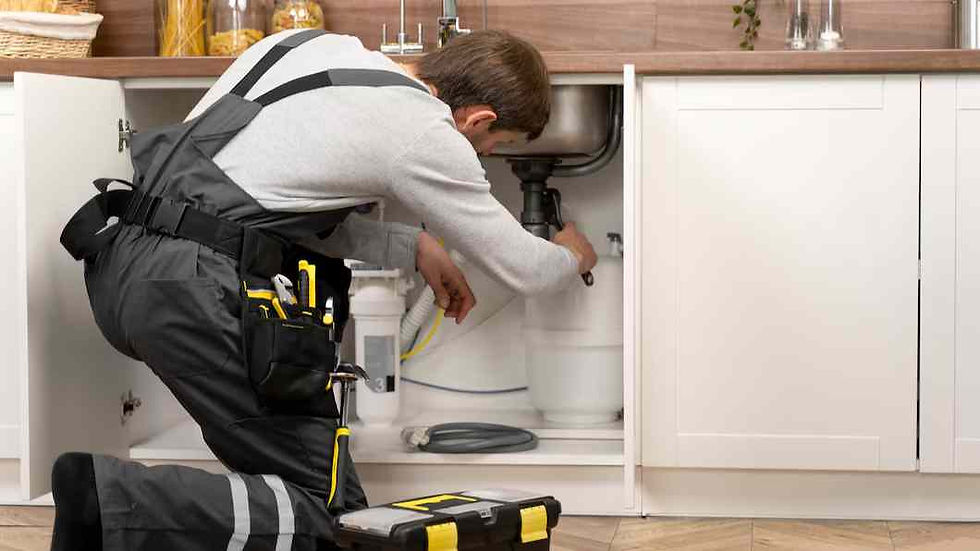 A plumber in black overalls fixing a pipe under a kitchen sink. White cabinets, tools, and a tool bag are visible. Bright and organized setting.