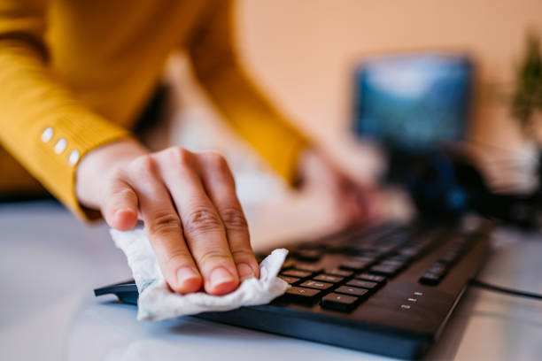 Hand in yellow sleeve wiping keyboard with cloth on desk. Blurred background with computer screen. Cleanliness and hygiene focus.