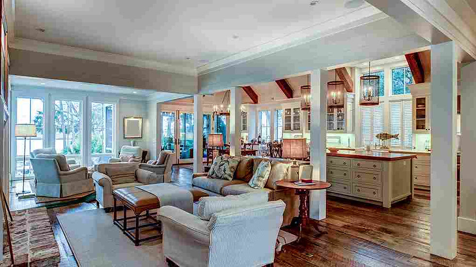 Spacious living room with beige sofas, armchairs, and a wooden floor. Open kitchen with pendant lights, warm lighting, and large windows.