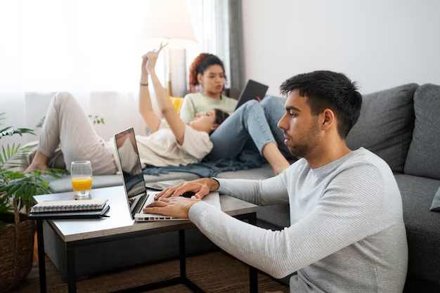 Man using laptop on table, woman reading on tablet, child playing on couch. Living room with gray sofa, notebook, and orange juice.