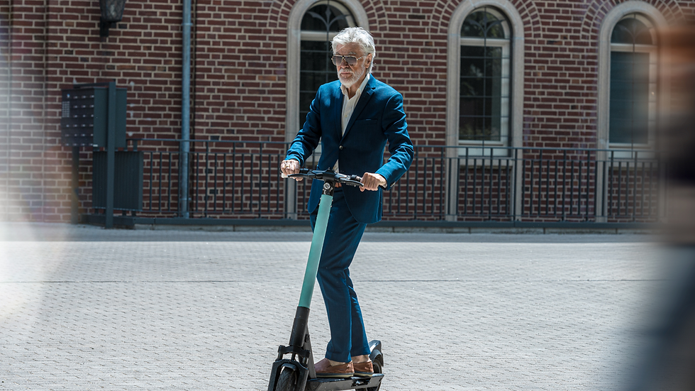 Man in a blue suit rides a teal scooter on a cobblestone street. Brick building with arched windows in the background. Calm and focused.