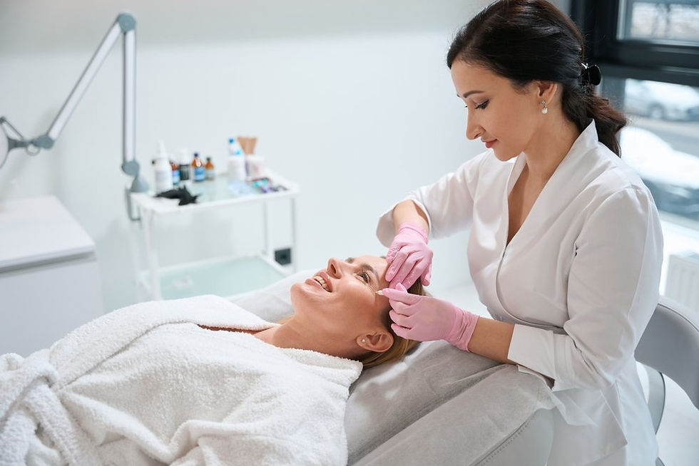 Aesthetician in white coat and pink gloves attends to a smiling client in a spa. Background has skincare products on a trolley.
