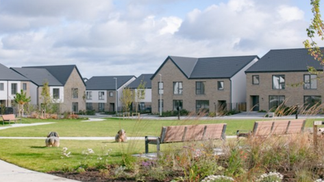 Cairn, Electrical Fit-Out by Greenlight, Residential Development – Contemporary two-storey homes with a mix of light brick and cream render facades, set around a landscaped communal green with benches, pathways, and planted flower beds.