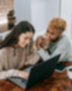 Two young people working on a laptop at a table, one supporting the other.