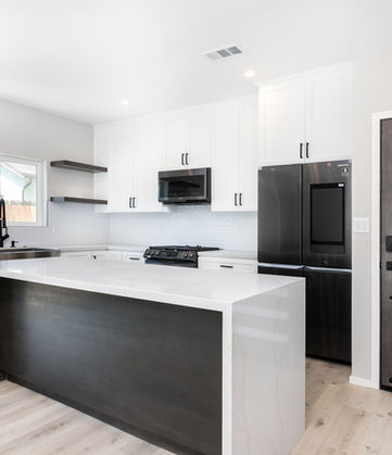 Modern kitchen with a large waterfall island, white cabinets, black appliances, floating shelves, and a dark front door with frosted horizontal panels.