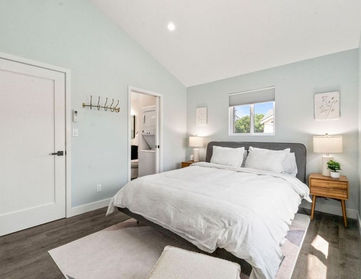 Staged primary bedroom with vaulted ceiling, soft light-colored walls, wide-plank flooring, a large bed with white bedding, two nightstands with lamps, and a window letting in natural light.
