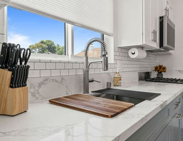 Close-up of a finished kitchen counter with marble-look quartz, white subway tile backsplash, deep sink with a tall pull-down faucet, and a window above the sink.
