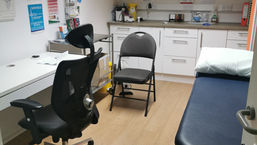 Medical office with chairs, examination table, and cabinets. Posters and brochures on the wall. Calm, organized setting with wood floors.