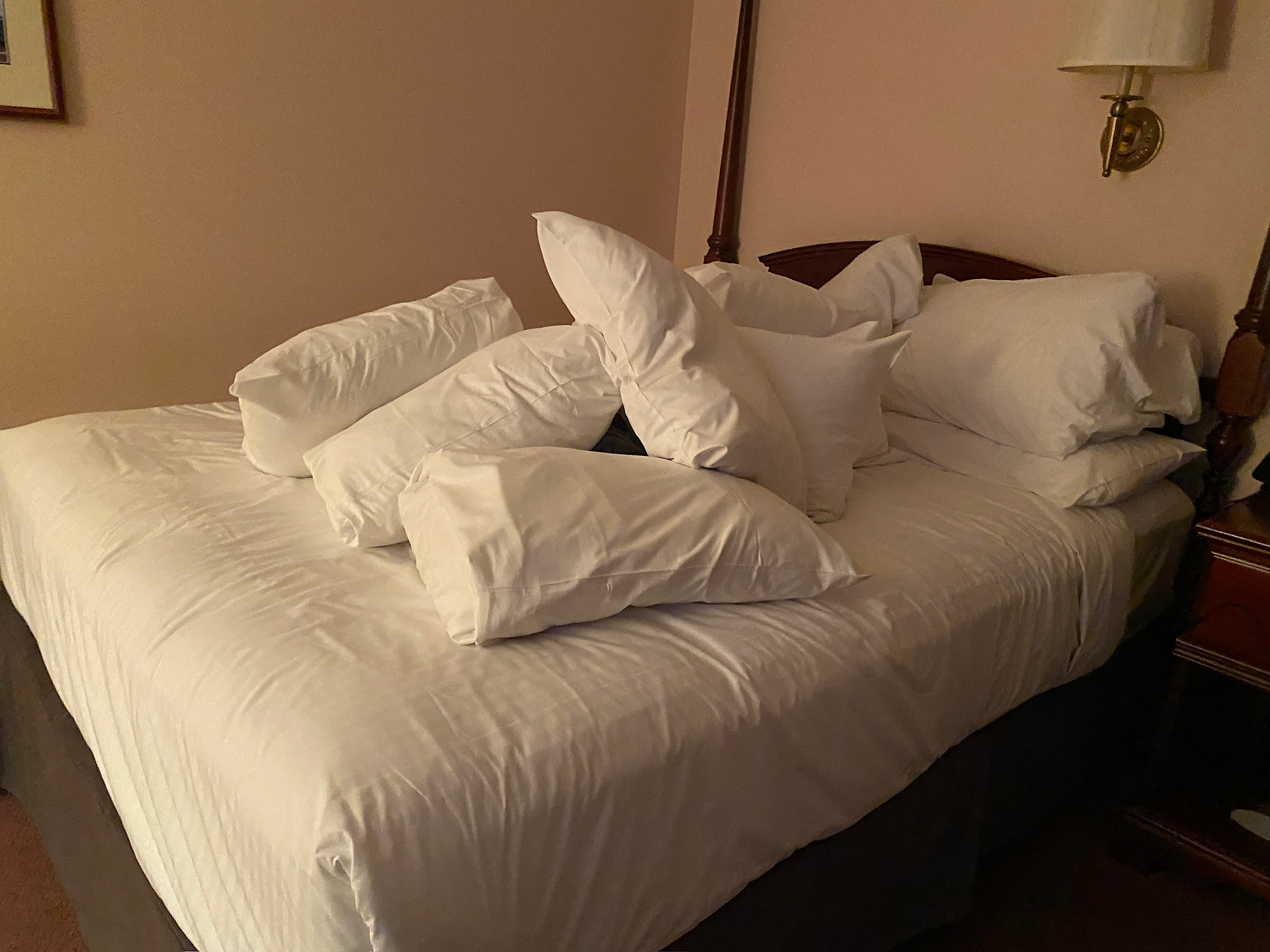 A hotel bed with a large pile of white pillows stacked haphazardly in the center, giving the impression of either a playful moment or an overwhelming abundance of pillows. The room has warm lighting, a wooden headboard, and a bedside lamp attached to the wall.