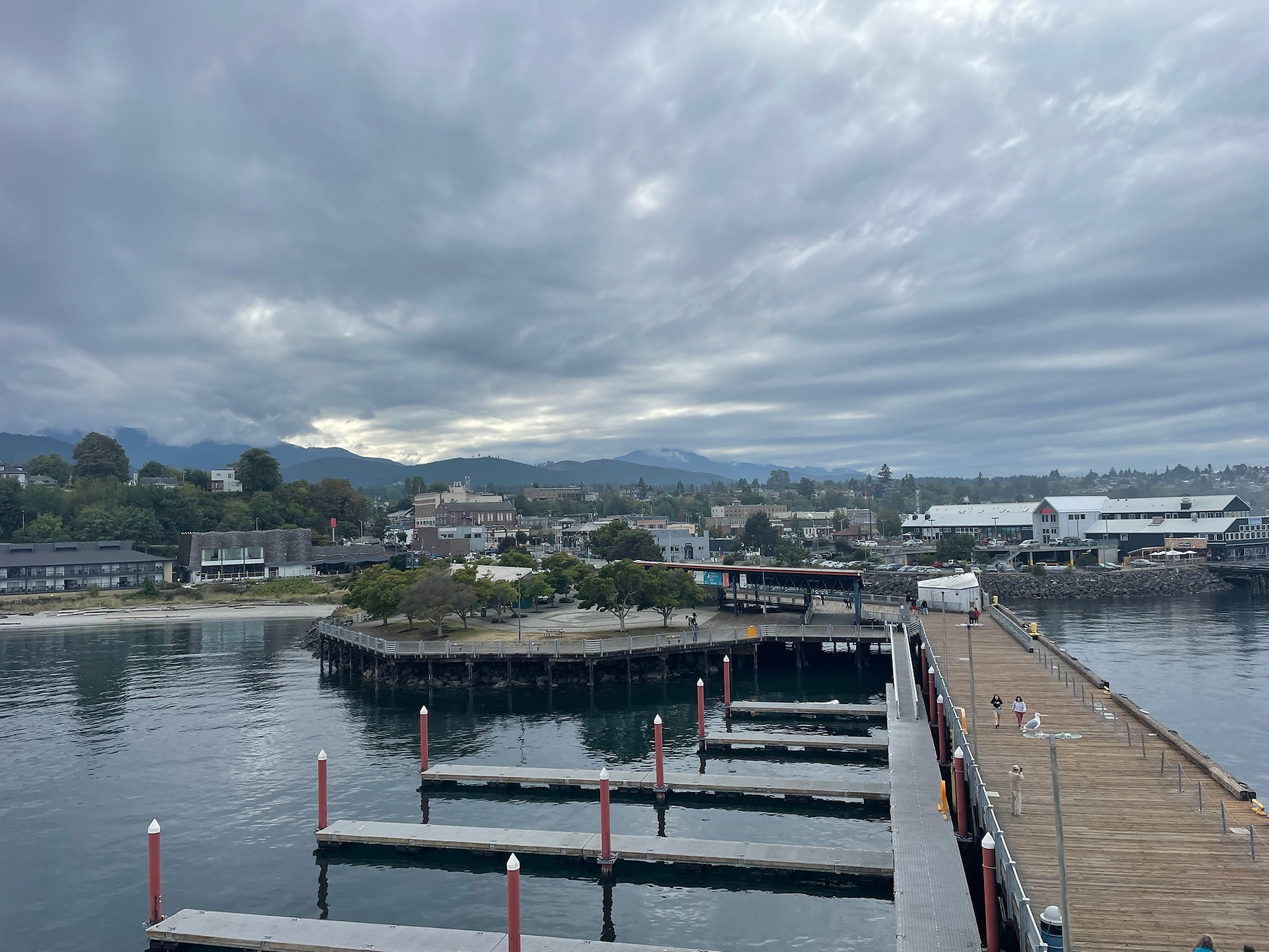 Cloudy morning view of Port Angeles, Washington, featuring the marina docks, wooden pier with people walking, and a backdrop of tree-lined buildings and misty Olympic Mountains.