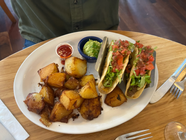 A breakfast plate at Blue Fox Café featuring two loaded breakfast tacos filled with scrambled eggs, lettuce, tomato, and salsa, served with golden crispy breakfast potatoes, a dollop of guacamole, and a side of salsa.