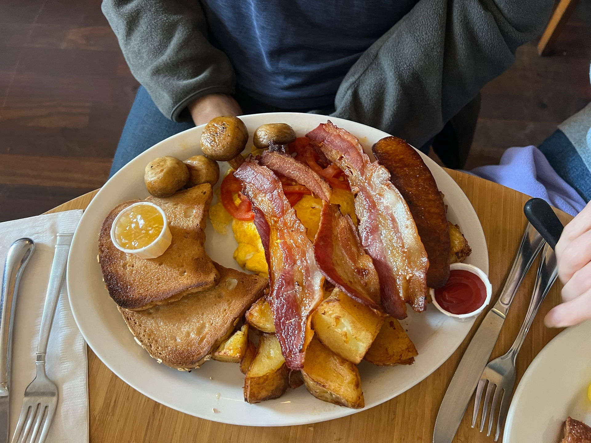 A hearty breakfast plate at Blue Fox Café featuring crispy bacon strips, scrambled eggs, grilled mushrooms, golden breakfast potatoes, toast with a pat of jam, and a slice of sausage—all artfully arranged and served with a side of ketchup.