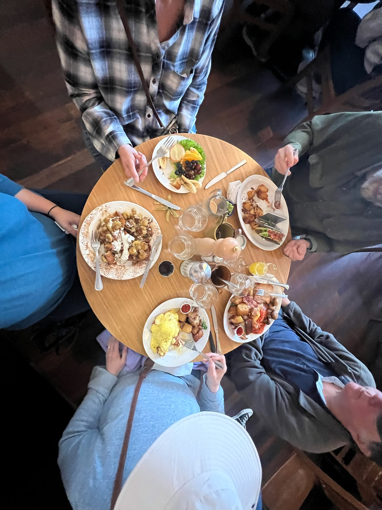Top-down view of a round wooden table at Blue Fox Café with five people enjoying a hearty breakfast. Plates are filled with omelets, breakfast potatoes, fruit, pancakes, and toast. A white hat and various colorful outfits add a casual, friendly vibe. Glasses of water and mugs of coffee are placed around the table.Top-down view of a round wooden table at Blue Fox Café with five people enjoying a hearty breakfast. Plates are filled with omelets, breakfast potatoes, fruit, pancakes, and toast. A white hat and various colorful outfits add a casual, friendly vibe. Glasses of water and mugs of coffee are placed around the table.