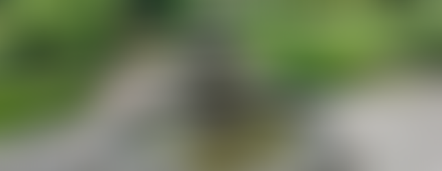 Stone tiered fountain surrounded by a circular basin, gently flowing water, and a peaceful garden path with wooden benches and blooming rose bushes in the background.