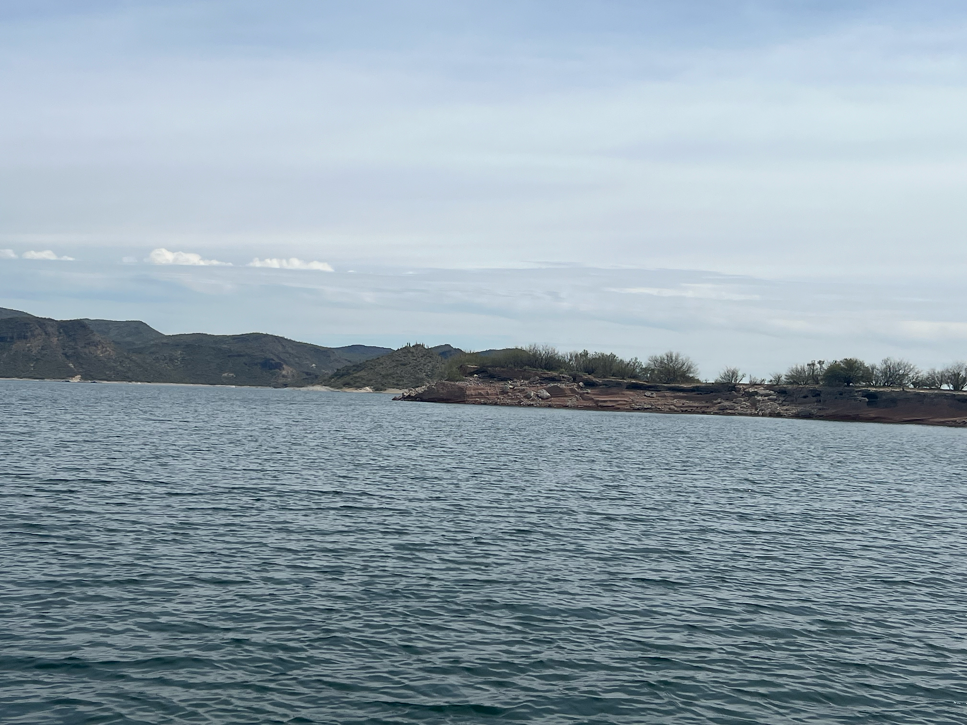 Wide view of Lake Pleasant with calm waters stretching to distant rolling hills and a rocky, shrub-covered shoreline under a lightly clouded sky.