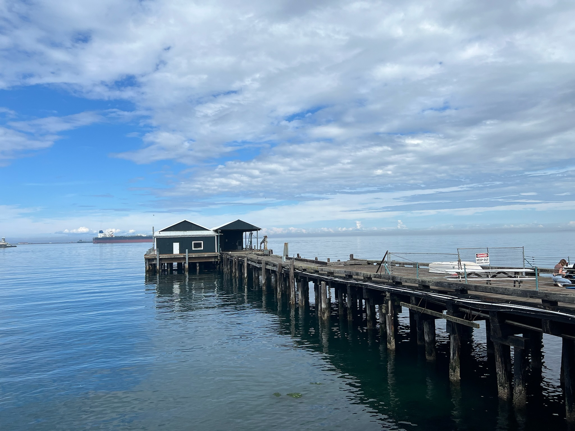 Wooden pier with two small black buildings extending over calm blue water in Port Angeles, Washington, with a large cargo ship visible in the distance under a partly cloudy sky.