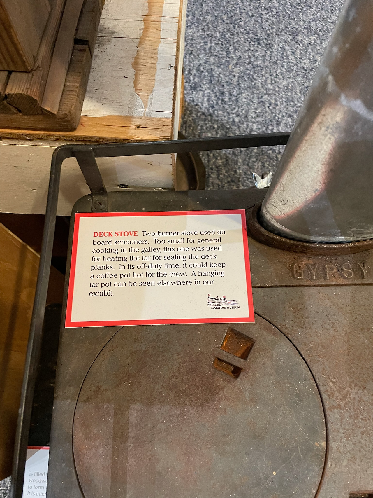 Close-up of an old two-burner deck stove on display at the Poulsbo Maritime Museum. The stove, labeled “GYPSY,” is rusty and well-worn, with an informational placard explaining that it was used on board schooners to heat tar for sealing deck planks, and occasionally to keep coffee warm for the crew. The card notes the stove was too small for general galley cooking.
