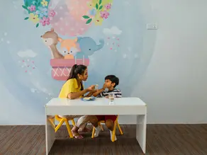 Smilestones therapist feeds a young boy at a white table in a playful pastel room with cartoon animals and a hot-air balloon mural.