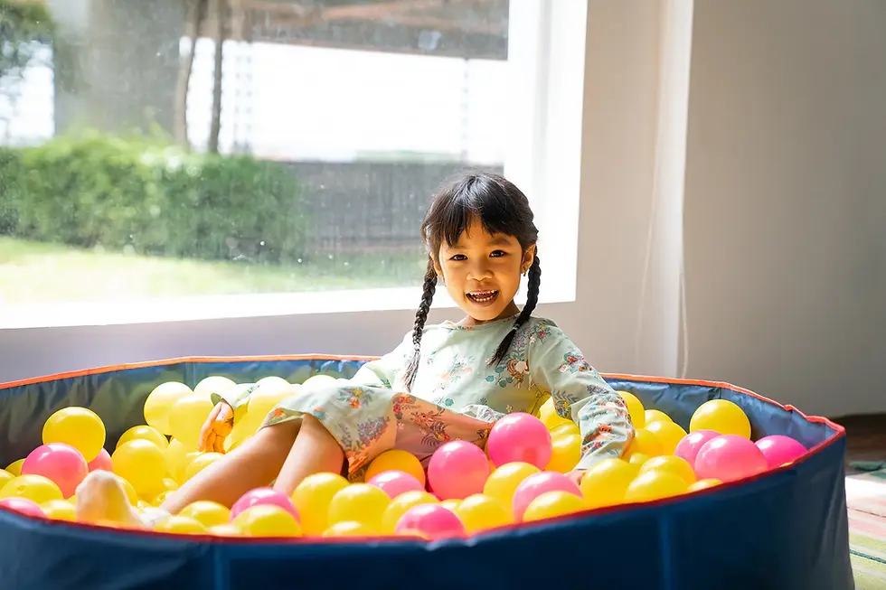 Smiling Khmer girl with braids sits in a blue ball pit filled with yellow and pink balls beside a bright window indoors at Smilestones Cambodia
