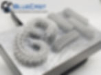 3D printed, intricate white resin design on a metallic surface. "BlueCast High Performance Castable Resin" . X-One V2
