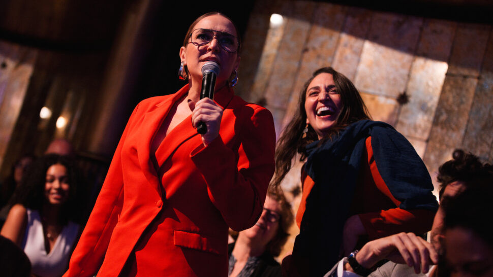 Jessica Vosk and Sara Bareilles stand together in the audience of Amplify. Sara is laughing. Jessica has a microphone.