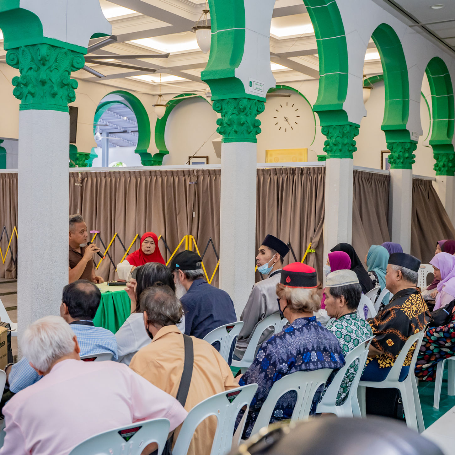A group of elderly people, mostly in traditional clothing, sit on plastic chairs listening to a speaker at the front of a hall with green and white Islamic architectural features.